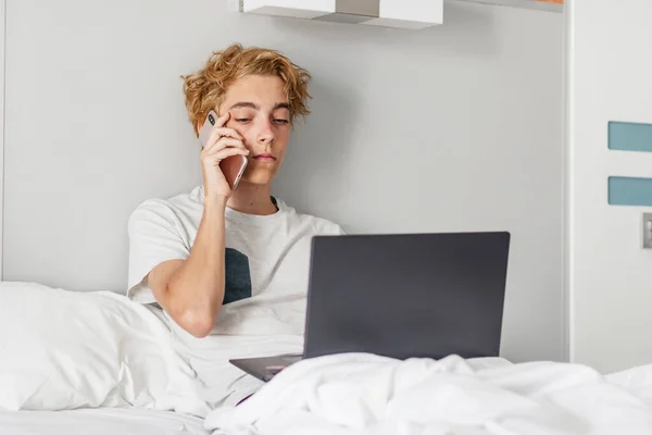 英俊的白人商人坐在酒店的床上 用手机聊天 有智能手机和手提电脑的青少年 — 图库照片