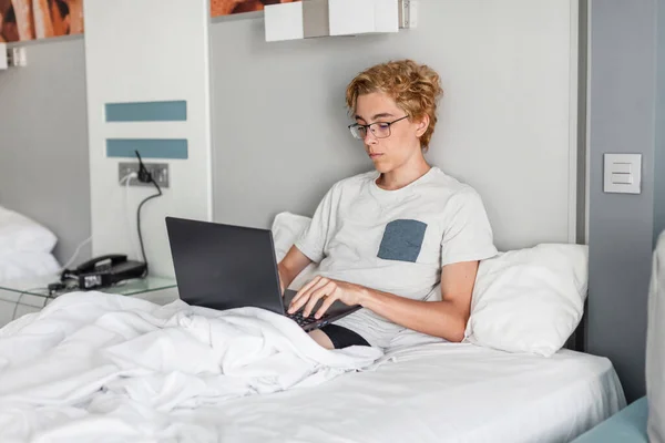 英俊的年轻人20多岁 穿着基本的灰色T恤 躺在笔记本电脑上 床上铺着带条纹的枕毛毯 悠闲地呆在卧室里 在校园或热点地区带着笔记本电脑的十几岁男孩 — 图库照片