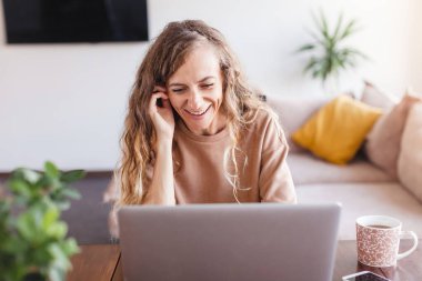 Oturma odasındaki kanepede otururken gümüş dizüstü bilgisayar kullanan neşeli bir kadın.