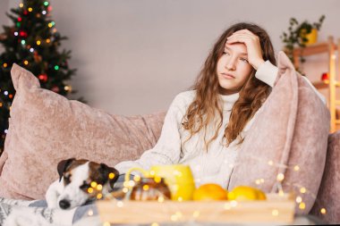 Evdeki telefona bakan üzgün genç bir kadın. Yalnız başına üzgün, cep telefonlu bir genç kız bir Noel ağacına karşı oturma odasında oturuyor.