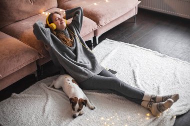 Odadaki kanepenin yanında oturmuş kulaklıkla müzik dinleyen neşeli genç bir kadın. Evdeki genç kız.