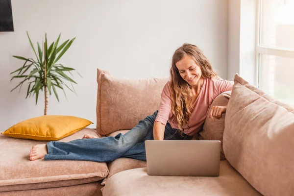 美しい陽気な女性を使用して銀のノートパソコンでソファの上に座っている間自宅で — ストック写真