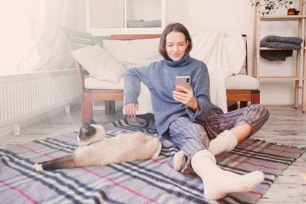 幸せな千年紀の白人の少女は自宅でスマートフォンを持っているソーシャルメディアをチェックします 猫と笑顔の若い女性は携帯電話アプリを使用してゲームをプレイ オンラインショッピング 注文配達はソファでリラックス — ストック写真