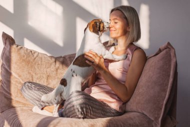 Orta yaşlı bir kadın hafta sonunu evde geçiriyor. Gülümseyen dişi, kanepede otururken sevgili köpeğiyle oynar.