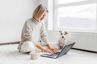 Köpekli mutlu orta yaşlı kadın rahat beyaz evinde dizüstü bilgisayarını kullanıyor. Oturma odasında çalışan serbest çalışan bir kadın.
