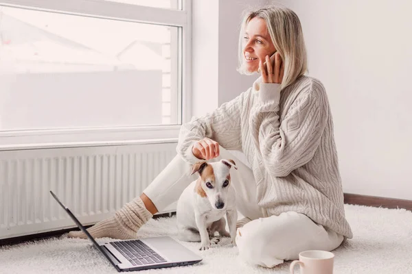 リラックスした中年の女性は彼女の家でノートパソコンや電話で働いています 大人の女性あります座っています床の近くにラジエーターと携帯電話でホワイトニットセーターで彼女の犬と離れて見て — ストック写真