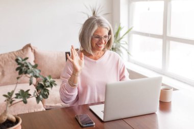 Bilgisayarda video görüşmesi yapan mutlu orta yaşlı kadın, ekrana el sallıyor, uzaktaki insanlarla sohbet ediyor. Gülümseyen, gri saçlı, son sınıf öğrencisi bir kadının, evdeki oturma odasındaki bir video görüşmesinde el sallarken görüntüsü.