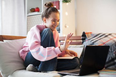 Mesafe dersi başlıyor. Evdeki kanepede oturan genç bayan öğrenci. Gülümseyen kız öğrenci dizüstü bilgisayara bakıp el sallıyor. Gençler video çağrısı ile iletişim kurar