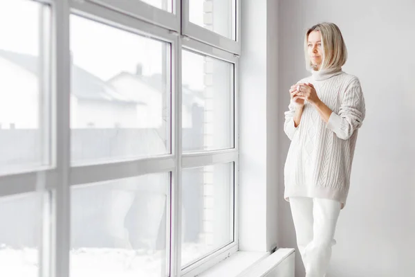 白い居心地の良いニットのセーターに身を包んだスタイリッシュな40代の女性の肖像画朝のコーヒーを楽しみ マグカップを持ち パノラマの窓を見ている女性 ロイヤリティフリーのストック写真