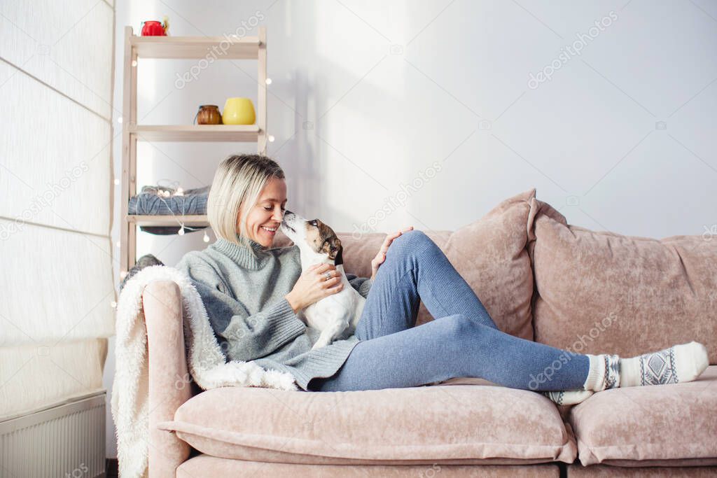Serene middle-aged lady housewife lounge sit on sofa and play with dog, calm 40s adult woman rest on comfort couch, puppy kissing female