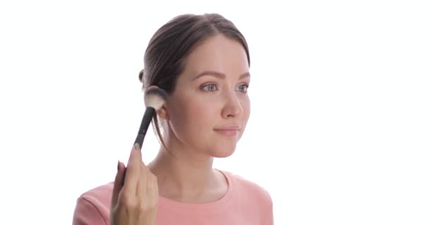 Porträtt av ung europeisk kvinna som gör rouge eller puder i ansiktet med sminkborste. Kvinna med ljusgröna ögon gör makeup på den vita bakgrunden. — Stockvideo