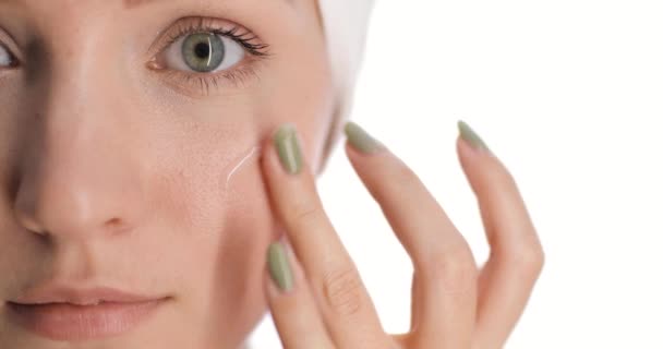 Primer plano de una parte de la cara de las mujeres jóvenes europeas con una toalla de secado de pelo en la cabeza aplica crema para el cuidado de la piel en la mejilla por su mano golpeando y sonriendo. — Vídeos de Stock