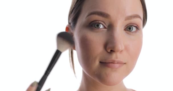 Close-up van jonge blanke vrouw die bloost op het gezicht met make-up borstel. Vrouw met mooie lichte ogen maakt make-up op de witte achtergrond. — Stockvideo