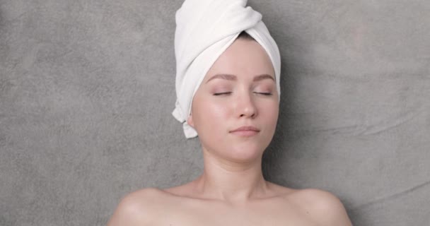 Porträt einer schönen jungen Frau mit einem Handtuch auf dem Kopf, die sich vor dem grauen Hintergrund zwei Gurkenscheiben über die Augen legt. — Stockvideo
