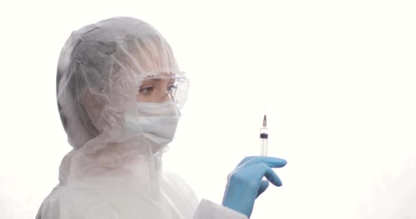 Médico con un traje protector con una jeringa que mira a una jeringa con vacuna, sobre el fondo blanco. Vista lateral. — Vídeos de Stock