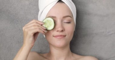 Kafasında havluyla gülümseyen güzel bir kadının portresi. Gri bir arka plana salatalık dilimleri koyuyor..