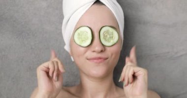 Kafasında havlu ve gri arka planda salatalık dilimleri olan güzel, pozitif ve güzel bir kadının portresi. Müzikle rahatlıyor..