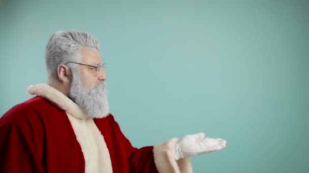 Bärtiger Mann in Nikolauskleidung lächelt und pustet Schnee in die Kamera, isoliert vor blauem Hintergrund - Weihnachtskonzept in Nahaufnahme — Stockvideo