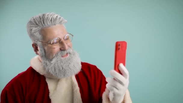 Zeitlupe des modernen Weihnachtsmannes telefoniert, per Videoanruf, aus der Ferne grüßt er, wünscht frohe Weihnachten. Schnee ins Telefon blasen. blauer Hintergrund. — Stockvideo