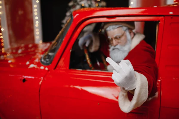 Angry, displeased, annoyed, stylish Santa Claus driving a red car. Santa Claus is preparing for Christmas. Shows the middle finger. Fuck sign.