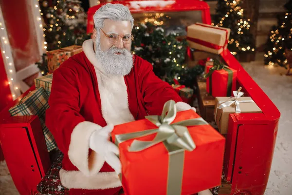 Πορτρέτο του ένα κομψό Santa Claus με ένα δώρο στα χέρια του, πολλά δώρα στο πίσω μέρος ενός αυτοκινήτου. Προετοιμασία δώρων για τον εορτασμό των Χριστουγέννων και της Πρωτοχρονιάς — Φωτογραφία Αρχείου
