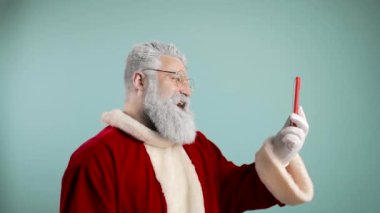 Modern Noel Baba 'nın yavaş çekim videosu telefonda konuşuyor, uzaktan, merhaba diyor, mutlu Noeller diliyor. Telefona kar üflemek. mavi arkaplan.