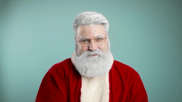 Vousatý muž v Santa Claus oblečení s úsměvem a foukání sníh do kamery, izolované přes modré pozadí - vánoční koncept zblízka — Stock video