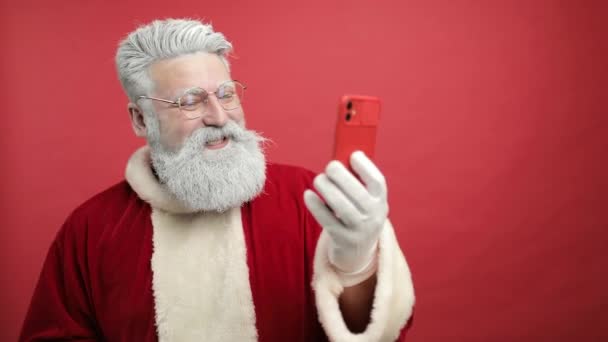 Babbo Natale moderno. Allegro Babbo Natale sta parlando al telefono, tramite videochiamata, a distanza, ti saluta, ti augura buon Natale. soffiando neve nel telefono. sfondo rosso. rallentatore — Video Stock