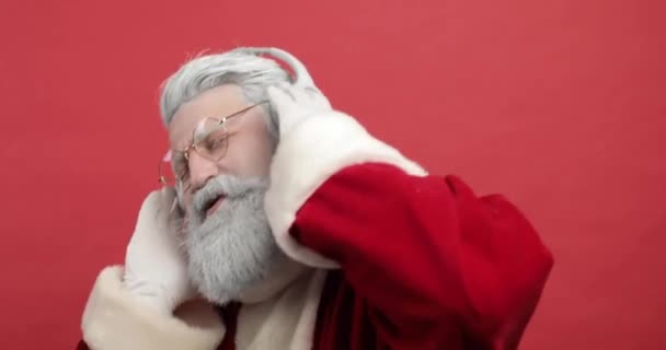 Der trendige, fröhliche Weihnachtsmann hört über Kopfhörer Musik, singt und tanzt auf rotem Grund. Weihnachtsstimmung, Weihnachtsfeier hautnah — Stockvideo