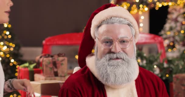 Una giovane donna sussurra qualcosa all'orecchio di un Babbo Natale sorpreso seduto in uno studio di Capodanno con le luci, accanto a un camion rosso con regali di Capodanno. — Video Stock