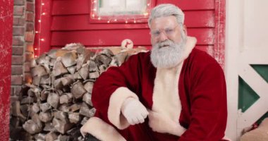 Gözlüklü, modern, yakışıklı Noel Baba 'nın portresi güzel bir yeni yıl dekore edilmiş kırmızı evin arka planında, ışıklar ve odun yığınıyla..