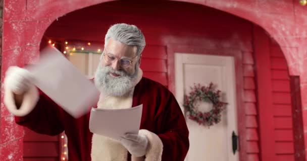 Potret Santa Claus, dengan luar biasa dan tertawa membaca surat-surat dalam Tahun Baru yang indah dihiasi studio dengan latar belakang rumah merah dengan lampu. — Stok Video