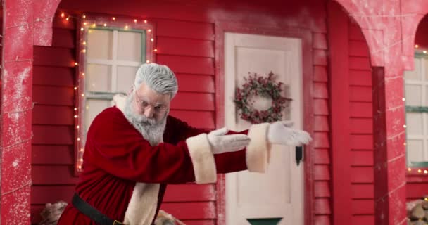 Activo Santa Claus invita a alguna parte, haciendo gestos de la mano contra el fondo de una casa roja con luces en un hermoso estudio decorado Año Nuevo. — Vídeo de stock