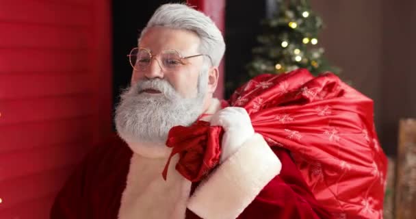 Sympatický Santa Claus s velkým pytlem dárků kráčí až na Nový rok zdobené červený dům se světly. — Stock video