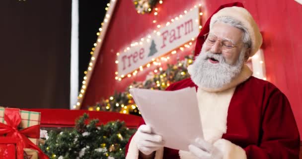 Портрет милого радостного Санта-Клауса, читающего письма в красивой новогодней студии. Улыбаясь и смеясь. — стоковое видео