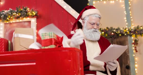 Portrait of a disgruntled and angry Santa Claus reading letters in a beautiful New Years decorated studio. He gets angry, throws away and tears up some of the letters. — Stock Video