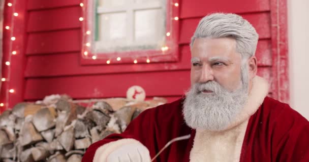 Un retrato de un guapo Santa Claus moderno abriendo sus gafas sobre el fondo de una hermosa casa roja decorada de Año Nuevo, con luces y pila de madera. — Vídeos de Stock