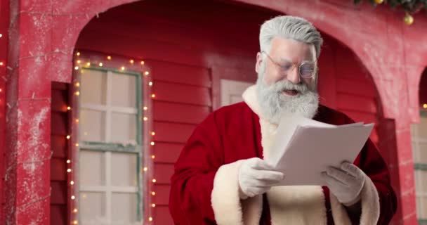 Portrait of a Santa Claus amazedly reading letters in a beautiful New Years decorated studio on the background a red house with lights. — Stock Video