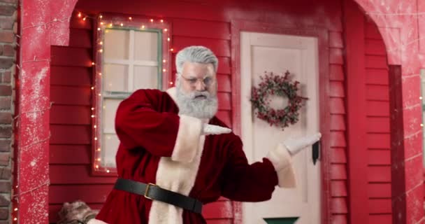 Σύγχρονη Santa Claus είναι αστείο χορό στο παρασκήνιο ένα κόκκινο σπίτι με φώτα σε ένα όμορφο Πρωτοχρονιάτικο διακοσμημένο στούντιο. Χαμογελώντας. — Αρχείο Βίντεο