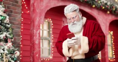 Modern Noel Baba arka planda akıllı telefonuyla bir oyun oynuyor güzel bir Yeni Yıl stüdyosunda kırmızı bir ev ışıklarla ve Noel ağacıyla süslenmiş. Kazandı..