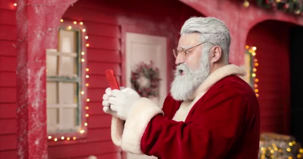 O Papai Noel moderno está jogando um jogo em um smartphone em segundo plano uma casa vermelha em um belo estúdio de decoração de Ano Novo com luzes e árvore de Natal. O quê? Vitória. Ele ganhou.. — Vídeo de Stock