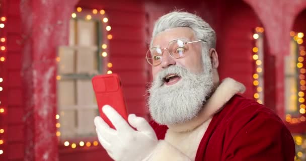Närbild av jultomten, leende och kommunicera via videolänk med någon som använder smartphone, på bakgrunden ett rött hus i en vacker nyår dekorerad studio. — Stockvideo