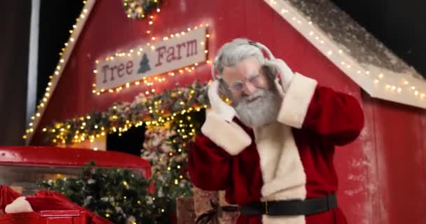 Santa Claus is listening to music with headphones, singing and dancing, on the background a red house in a beautiful New Years decorated studio. — Stock Video