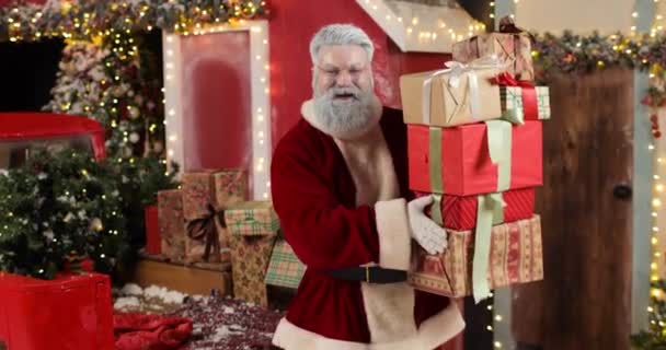 Santa Claus is dancing with a stack of boxes with gifts, smiling and looking at the camera, on Christmas Eve or New Year Eve. Beautiful New Years decorated studio. — Stock Video