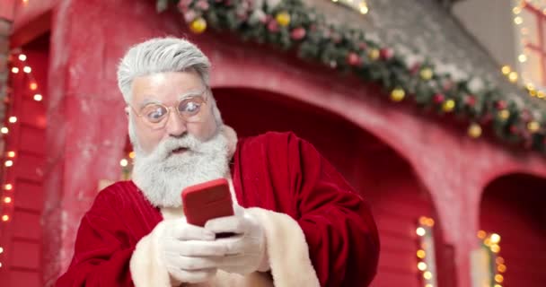 Σύγχρονη Santa Claus παίζει ένα παιχνίδι σε ένα smartphone στο παρασκήνιο ένα κόκκινο σπίτι σε ένα όμορφο Πρωτοχρονιάτικο διακοσμημένο στούντιο με φώτα και χριστουγεννιάτικο δέντρο. - Ναι. Νίκη. Κέρδισε.. — Αρχείο Βίντεο
