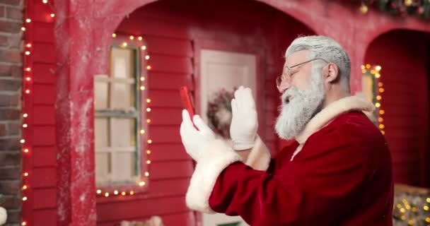 Санта-Клаус общается по видеосвязи с кем-то с помощью смартфона на фоне красного дома в красивой новогодней студии. Вид сбоку. — стоковое видео