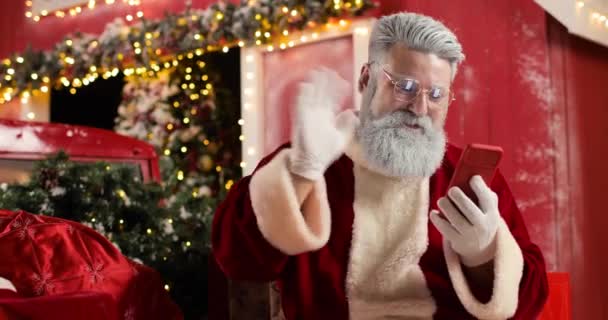 Retrato de Santa Claus riendo y comunicándose por video usando smartphone, saludando a alguien, en el fondo una casa roja en un hermoso estudio decorado en Año Nuevo. — Vídeo de stock