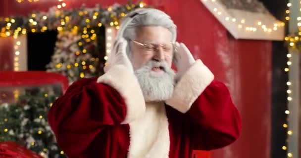 Portret Świętego Mikołaja słuchającego muzyki ze słuchawkami i śpiewającego na tle czerwonego domu w pięknym noworocznym studio. — Wideo stockowe