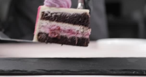 Mãos de um chef de pastelaria colocando um pedaço de bolo de chocolate enfeitado com mirtilo em um substrato preto. Vista frontal. — Vídeo de Stock