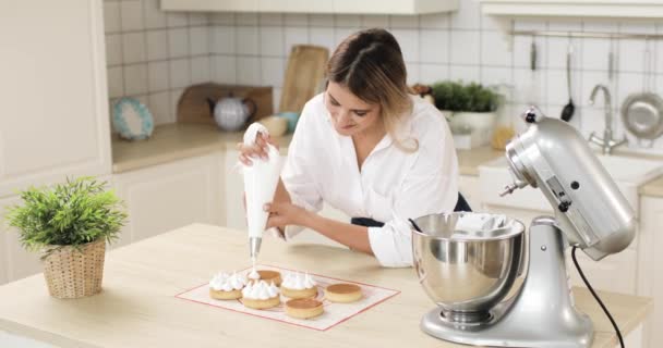 Une jeune pâtissière décore un tartelette aux biscuits avec de la crème tirée d'un sac à pâtisserie. Faire des gâteaux dans une pâtisserie maison. — Video